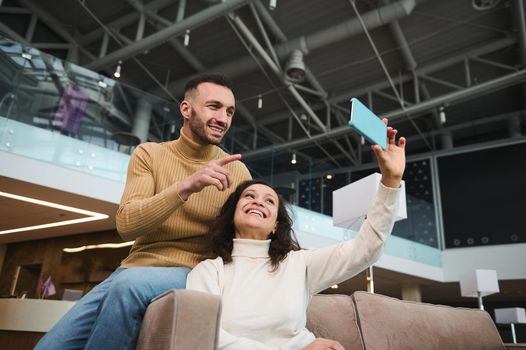 Handsome young multi ethnic couple in casual wear holding smartphone and looking at webcam talking via video call while relaxing in vip lounge waiting to board flight at international airport