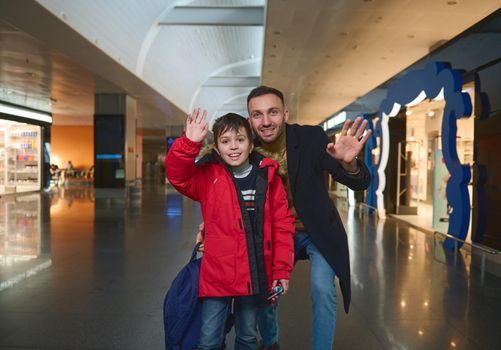 Happy Caucasian family of young cheerful dad and cute boy son waving looking at camera while standing outside duty free shops in the international airport departure terminal. Family air travel concept