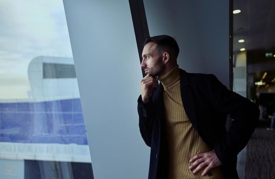 Charming thoughtful Middle Eastern man transit passenger looking through panoramic windows overlooking runway at planes in the departure terminal of international airport waiting to board flight