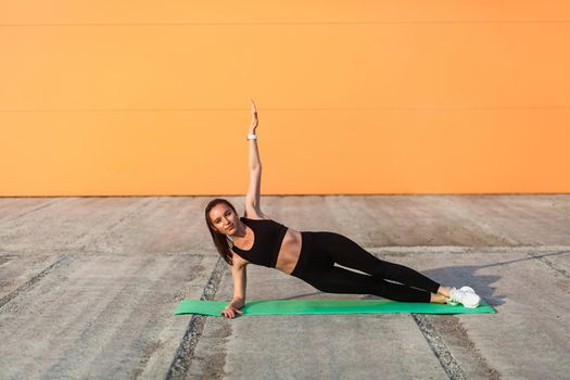 Athletic beautiful girl in tight sportswear, black pants and top, practicing yoga, doing Vasisthasana side plank pose with one hand, training muscles and strength. Health care, sport activity outdoor