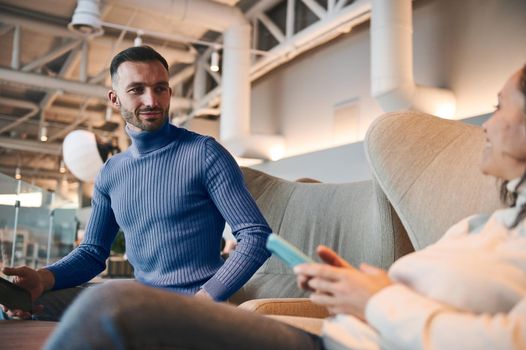 Attractive handsome Middle-Eastern young man, passenger in blue pullover relaxing with his girlfriend on the blurred foreground in the VIP lounge of the departure terminal of International airport