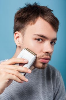 Portrait of handsome brunette man with bristle and mustache in casual sweater removing facial hair using epilator for smooth skin, male beauty care concept. studio shot isolated on blue background
