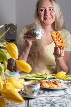 blonde middle-aged hugging a kitten and drinking tea, still life with yellow tulips and cakes a stack of books on the table. High quality photo