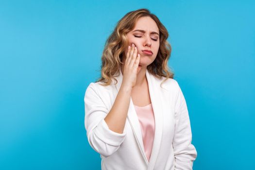 Toothache. Portrait of unhappy sick woman with wavy hair in white jacket suffering dental pain, standing with closed eyes and touching cheek to relieve pain. studio shot isolated on blue background