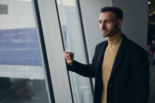 Portrait of charming handsome thoughtful Middle Eastern man, transit passenger standing at panoramic windows overlooking runway in the international airport departure terminal, waiting to board flight