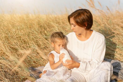 Little girl with her mother drink milk plastic bottle. Happy family, daughter with mom resting outdoor drink healthy diary product. Countryside rural scene. Caucasian female on nature