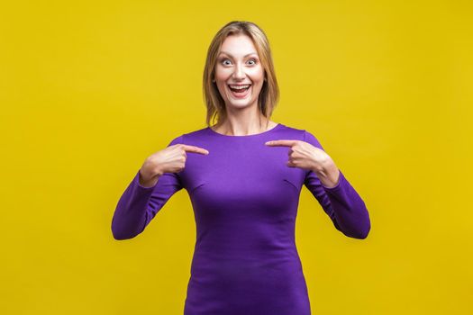 Portrait of joyous winner, excited businesswoman in elegant purple dress smiling broadly and pointing at herself, boasting of achievements. indoor studio shot isolated on yellow background
