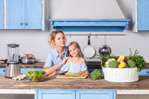 Young woman and lovely girl sitting in modern kitchen, looking at camera, mother and daughter cooking salad together, vegetarian food, fresh green vegetables and blender on table, healthy nutrition