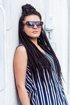 Portrait of young brunette woman with black afro american dreadlocks hairstyle in striped dress and sunglasses, standing, posing and looking at camera. indoor studio shot on white wall.
