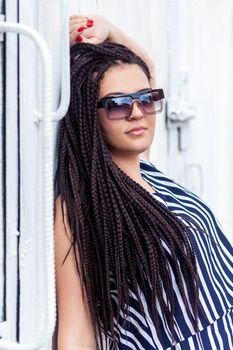 Portrait of young brunette woman with black afro american dreadlocks hairstyle in striped dress and sunglasses, standing, posing and looking at camera. indoor studio shot on white wall.