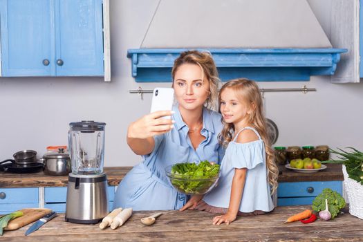 Charming woman and little girl taking selfie in modern kitchen, holding bowl of vegetable salad and posting photo in social network using cell phone, food blog about healthy nutrition, vegetarian diet