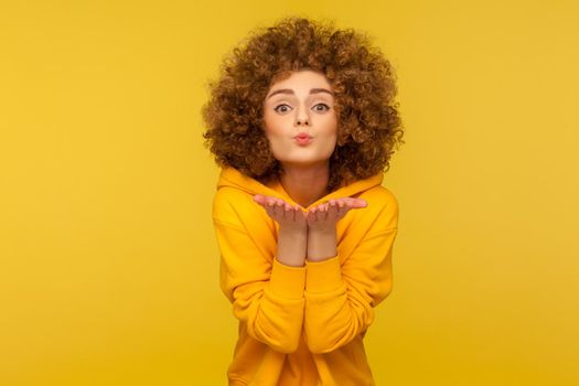 Catch my love kiss. Portrait of curly-haired young woman in urban style hoodie kissing air over palms, demonstrating romantic feelings, affection. indoor studio shot isolated on yellow background