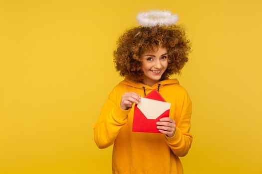 Love message. Portrait of happy curly-haired angelic woman with saint nimbus holding letter in envelope and looking at camera with playful smile. indoor studio shot isolated on yellow background