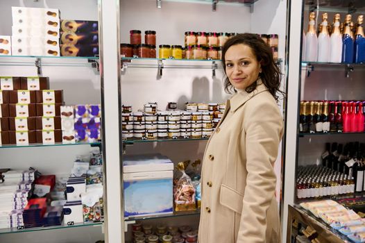 Attractive middle-aged multi-ethnic dark-haired elegant woman- transit passenger, traveler in casual beige coat looking at camera while shopping in the International Airport duty free shop