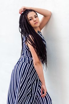 Portrait of serious young brunette woman with black afro american dreadlocks hairstyle in striped dress posing and looking at camera . indoor studio shot on white wall.