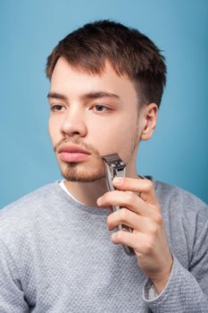 Grooming and self care. Portrait of concentrated focused brunette man shaving with trimmer or electric shaver, looking aside with serious attentive face. studio shot isolated on blue background