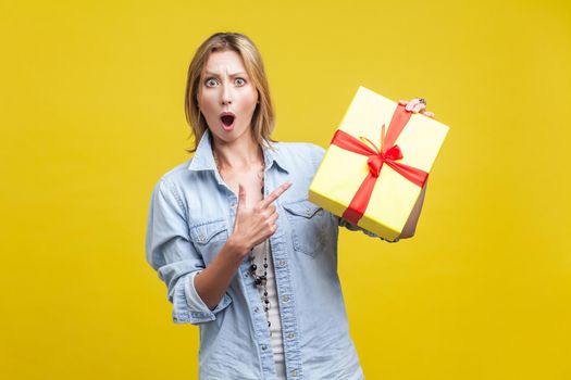 Portrait of amazed woman with fair hair in denim casual shirt pointing at wrapped gift box and looking with amazement, shocked by holiday present. studio shot isolated on yellow