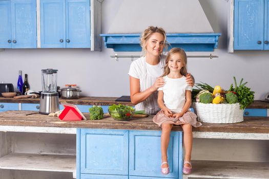 Beautiful mother in apron hugging daughter smiling at camera in kitchen with modern furniture, basket of fresh green vegetables and cooked salad on table, vegan healthy food, vegetarian nutrition