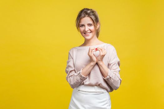 Portrait of pretty in love young woman with fair hair in casual beige blouse standing, showing heart sign with arms and looking at camera, romance. indoor studio shot isolated on yellow background
