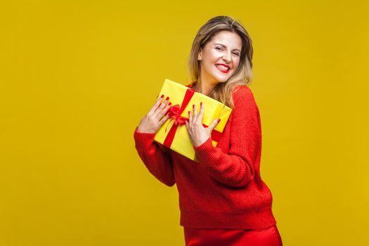 Portrait of extremely happy woman with red lipstick in bright casual sweater, holding wrapped gift box, smiling at camera, enjoying christmas present. indoor studio shot isolated on yellow background