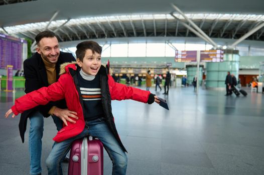 Adorable child boy with passport and boarding pass in his outstretched hands while his cheerful father is riding him on a suitcase in the departure hall of the airport, waiting check-in for a flight