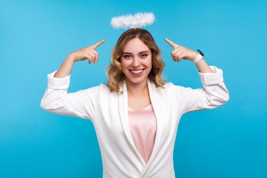 Portrait of attractive angelic woman with wavy hair in white clothes standing, pointing at halo on her head and smiling, cute sensual girl with nimbus. indoor studio shot isolated on blue background