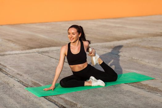 Smiling athletic girl in tight sportswear, black pants and top, practicing yoga, doing one-legged king pigeon pose, stretching leg muscles for better flexibility. Health care, sport activity outdoor