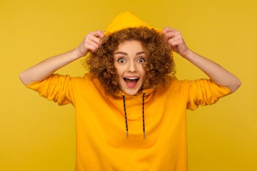 Portrait of funny amazed girl with curly hair wearing urban style hoodie, putting on hood and looking surprised at camera, youth sport style female fashion. studio shot isolated on yellow background
