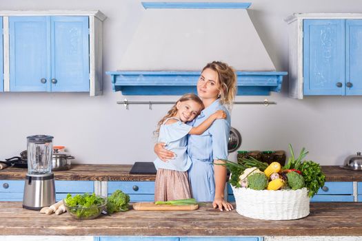Beautiful mother hugging lovely daughter and looking at camera, standing in kitchen with modern furniture, basket of fresh green vegetables and blender on table, vegetarian food, healthy nutrition