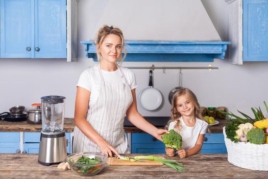 Cheerful pretty mother in apron and little daughter holding broccoli and smiling at camera, cooking salad in modern kitchen together, preparing vegetarian food, basket of fresh vegetables on table