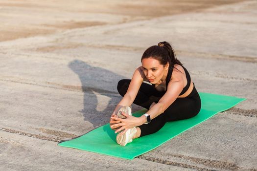 Positive athletic fit woman in tight sportswear, black pants and top, practicing yoga, doing Head-to-Knee Pose, touching toes, stretching leg and back muscles. Health care, sport activity outdoor