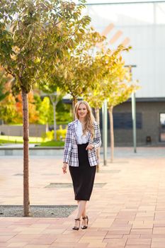 Full length portrait of stunning beautiful positive woman, confident businesslady with long blond hair wearing plaid jacket and skirt walking alone on sidewalk outdoors, office style clothes, fashion