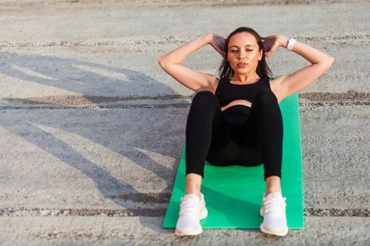 Athletic pretty girl in tight sportswear, black pants and top, lying on mat doing sit ups, crunch exercise, training abdominal muscles. Health care and weight loss concept, sport activity outdoor