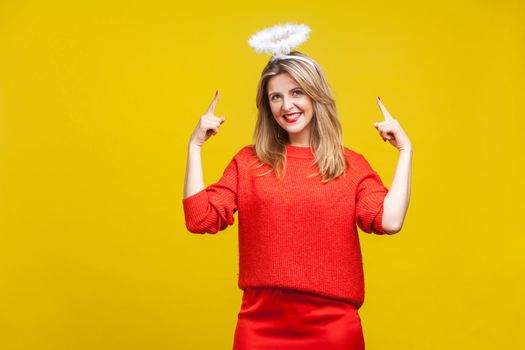 Portrait of proud adorable young woman with bright makeup in red casual clothes standing, pointing at hola on her head and smiling. indoor studio shot isolated on yellow background