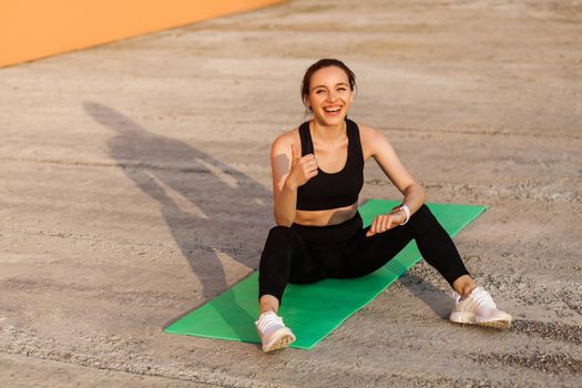 Delighted athletic girl in black pants and top, smiling and showing thumbs up, sitting on yoga mat after workouts, gesturing like, satisfied with physical training. Health care, sport activity outdoor