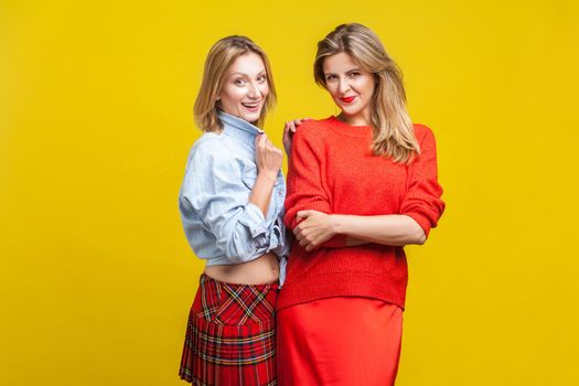 Best friends. Portrait of two fashion female models in stylish casual clothes standing together, smiling at camera. women showing autumn trends. indoor studio shot isolated on yellow background
