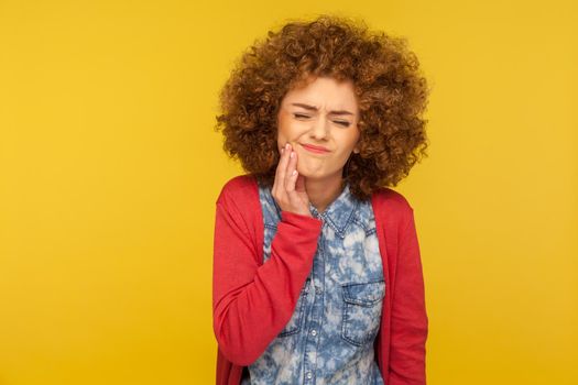 Dental problems. Portrait of unhealthy woman with curly hair wincing in pain and touching sore cheek, suffering unbearable toothache, gum disease. indoor studio shot isolated on yellow background