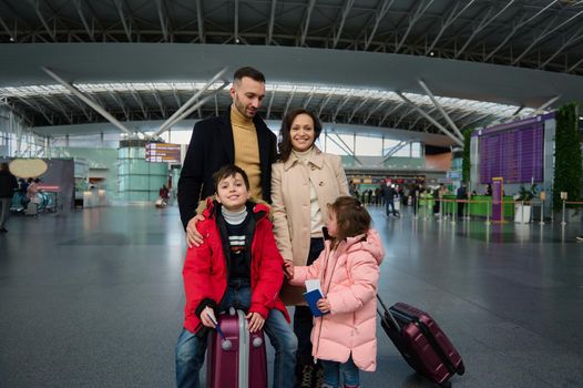 Happy family with children standing with suitcases in the departure hall of an international airport, waiting for flight check-in, customs control and boarding. Travel, journey, tourism, air travel