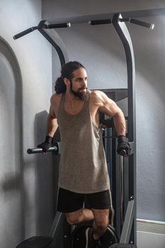 Full length portrait of confident young adult strong man with long curly hair working out in gym, hanging on horizontal bar with hands, doing pull ups with, exercises for triceps. indoor, looking away