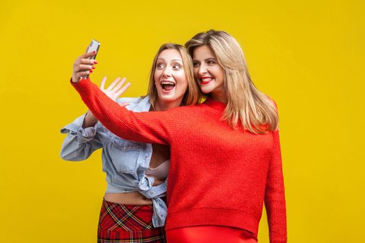 Best friends having fun. Portrait of happy women in stylish clothes standing with toothy smile and taking selfie on phone together, enjoying life. indoor studio shot isolated on yellow background
