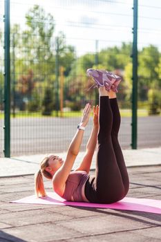 Motivated fit woman in tight pants lying on yoga mat and reaching hands to feet, training outdoor summer day, doing raised leg crunch, exercising abdominal muscles. Sports activity for weight loss