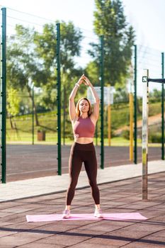 Fit athletic woman in tight pants practicing yoga on mat outdoor summer day, standing with raised hands and meditating, doing balancing asana, feeling peace and harmony. Health care, sports activity