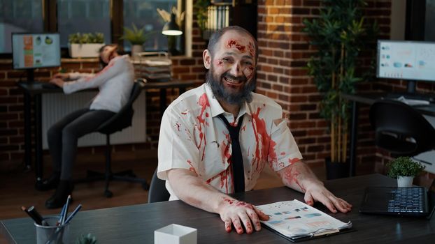 Creepy looking office zombie with bloody and deep face scars smirking spooky while giving thumbs up. Brain dead apocalyptic looking animated corpse smiling evil at camera while sitting in office.