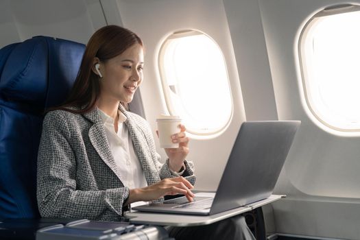 Attractive asian female passenger of airplane sitting in comfortable seat listening music in earphones while working at modern laptop computer