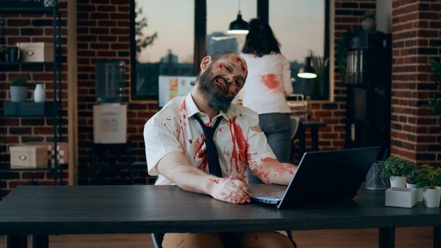 Mindless brain-eating zombie working on laptop while smirking creepy at camera. Dangerous brain dead monster with deep and bloody scars trying to use computer but fails big time.