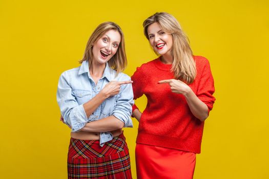 Best friends joking. Portrait of two happy women in casual clothes pointing and laughing at each other, looking at camera, carefree female friendship. indoor studio shot isolated on yellow background