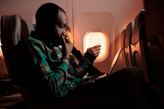 African american employee working on laptop during plane flight, travelling to international destination to go on work trip. Young man flying in economy class with airplane transport.