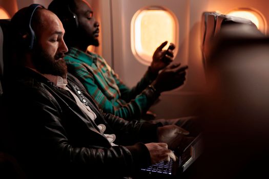 Multiethnic group of people flying in economy class on plane jet, travelling to holiday destination. Using laptop and smartphone during sunset flight before arriving on vacation trip.
