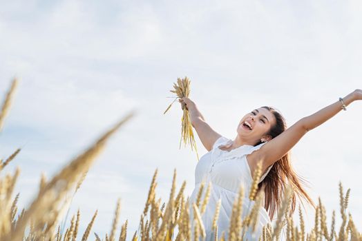 Young brunette woman standing in golden field holding heap of rye and wearing white dress lit by sunset light, laughing, copy space