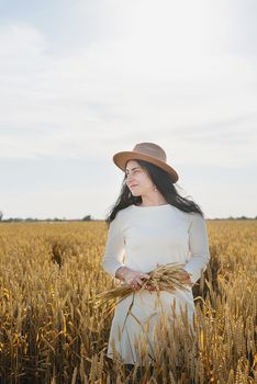 Mid adult woman in white dress walking and dancing across golden field holding heap of rye lit by sunset light, copy space